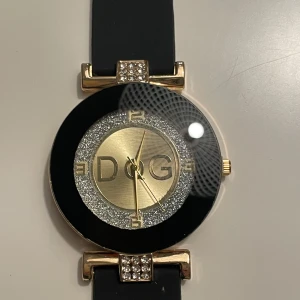 Svart och guld klocka från D&G - Elegant klocka från D&G med svart armband och en glittrig urtavla i guld och silver. Klockan har en rund boett med dekorativa stenar på fästena. Perfekt för att ge en lyxig touch till din stil. Replika!!!