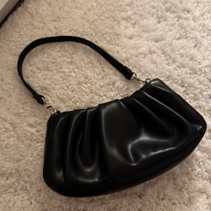 Svart axelväska i syntet - Snygg svart axelväska i syntetmaterial med dragkedja och silverfärgade detaljer. Aldrig använd så skicket är nytt, säljer för 200