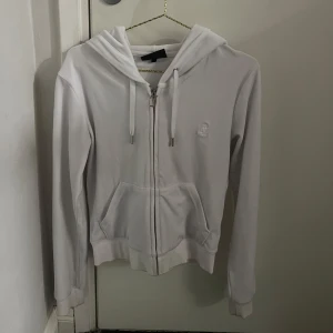 Vit hoodie från Juicy Couture - Säljer en stilren vit hoodie från Juicy Couture med dragkedja och huva. Den har en broderad logga på bröstet och praktiska fickor framtill. Perfekt för en avslappnad look.
