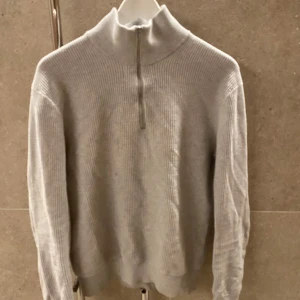 Grå ribbad tröja från East West - Säljer en stilren grå ribbad tröja från East West. Tröjan har en dragkedja vid halsen och långa ärmar, perfekt för kyligare dagar. Den är både bekväm och snygg, ett måste i garderoben! Säljes då den blivit förliten. Liten prick på tröjan på framsidan men annars fin och i bra skick