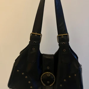 Svart axelväska med detaljer - Snygg svart axelväska i skinn med guldfärgade detaljer och en stor ring framtill. Väskan har dubbla axelremmar och dekorativa nitar som ger en cool look. Perfekt för att ge din outfit en edgy touch.