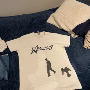 Vit t-shirt med tryck - Säljer en vit t-shirt från Gildan med ett coolt svart tryck av silhuetter och text. T-shirten är kortärmad och tillverkad i 100% bomull, vilket gör den bekväm att bära. Perfekt för en avslappnad stil.