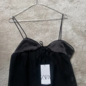 Svart topp från Zara - Säljer en elegant svart ärmlös topp från Zara med tunna axelband och knytning framtill. Toppen har en luftig design och är perfekt för varma dagar. Den är märkt med Zara och har en stilren look. Storlek M men den skulle passa xs-s skulle jag säga. Icke använd eftersom jag fick två av samma i paketet.