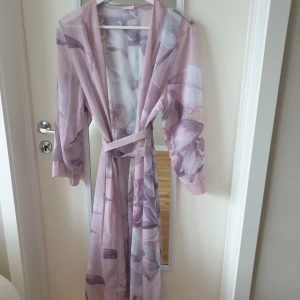 Ljusrosa blommig kimono från Oysho - Säljer en elegant ljusrosa kimono från Oysho med ett vackert blommigt mönster. Kimonon har vida ärmar och ett knytband i midjan. Perfekt för en avslappnad och stilfull look.