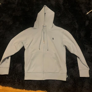 Turkos Ralph lauren hoodie med dragkedja - Säljer en stilren turkos hoodie från Polo Ralph Lauren. Den har en dragkedja framtill och en liten broderad logotyp på bröstet. Perfekt för en avslappnad stil med sina långa ärmar och justerbar huva.