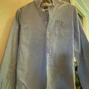 Blå skjorta från GANT - Säljer en stilren blå skjorta från GANT med klassisk krage och knappar framtill. Skjortan har en bröstficka med broderad logotyp och är långärmad. Perfekt för en snygg och avslappnad look. Skjortan är i mycket bra skick och har inga defekter. Pris kan diskuteras. 