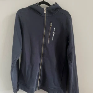 Mörkblå huvtröja från Sail Racing - Snygg mörkblå huvtröja från Sail Racing med dragkedja framtill och en praktisk bröstficka med logotyp. Tröjan har en bekväm huva och långa ärmar, perfekt för kyligare dagar. Använd fåtal gånger. 