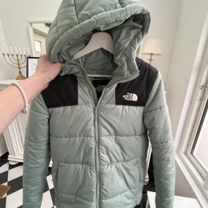 Grön pufferjacka från The North Face ❄️ - Snygg grön pufferjacka från The North Face med svarta detaljer på axlarna. Jackan har en praktisk huva och dragkedja framtill. Perfekt för kyligare dagar.