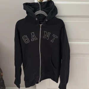 Svart hoodie från GANT - Säljer en svart hoodie från GANT med dragkedja och huva. Tröjan har GANT-loggan i glittriga stenar på framsidan och praktiska fickor. 🤗