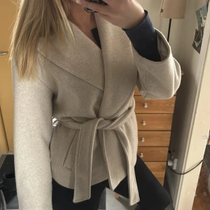 Beige kappa med knytband - Säljer en stilren beige kappa med knytband i midjan. Jackan har en omlott-design och långa ärmar, perfekt för kyligare dagar. En tidlös och elegant look som passar till många tillfällen.