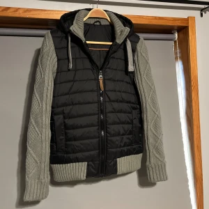 Cardigan jacka med stickade armar - Säljer en snygg svart och grå jacka med quiltad design och stickade ärmar. Jackan har en dragkedja framtill och en praktisk huva med dragsko. Perfekt för kyligare dagar!