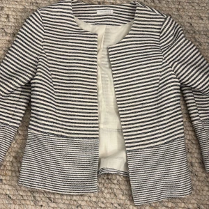 Randig kavaj från Promod - Snygg randig kavaj från Promod i svart och vit. Den har en stilren design utan knappar och är perfekt för en chic look. Kavajen har långa ärmar och en normal passform. Perfekt för att ge din outfit en elegant touch.