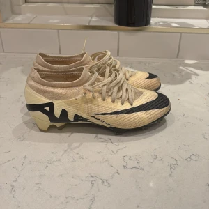 Beige och svarta fotbollsskor från Nike - Snygga fotbollsskor från Nike i beige med svarta detaljer och snörning. Skorna har en modern design med Nike-loggan tydligt synlig på sidan. Perfekta för fotbollsplanen med en tight passform.
