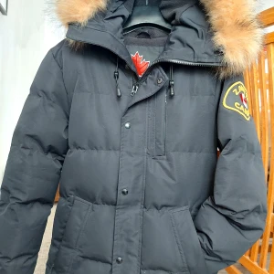 Svart parkajacka från Zavetti Canada - Säljer en svart parkajacka från Zavetti Canada med pälsfodrad huva och broderad logga på ärmen. Jackan har dragkedja och knappar framtill samt flera praktiska fickor. Perfekt för kalla dagar!