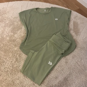 Grön träningstopp och leggings - Säljer ett set med en grön träningstopp och matchande leggings. Toppen har en lös passform och korta ärmar, perfekt för träning. Materialet är mjukt och stretchigt. Köpt från lager 157 o aldrig använd