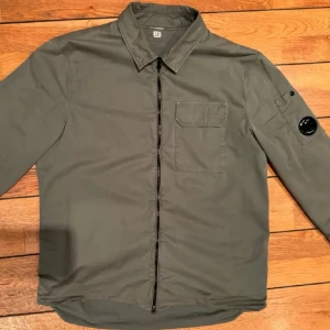 Grön overshirt med dragkedja - Snygg grön overshirt från C.P. Company med dragkedja framtill och en bröstficka. Den har en cool detalj med en knapp på ärmen. Perfekt för lager-på-lager-stil. Prutning ignoreras, skriv ifall intresse!