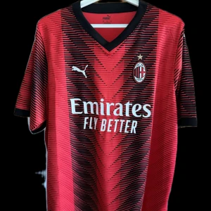 Röd AC Milan fotbollströja från Puma - Snygg röd AC Milan fotbollströja från Puma med svart och rött mönster. Tröjan har korta ärmar och klubbens emblem på bröstet. Perfekt för fans som vill visa sitt stöd.