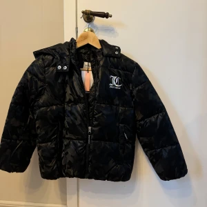 Svart pufferjacka från Juicy Couture - Säljer en svart pufferjacka från Juicy Couture med mönster och logga på bröstet. Jackan har en huva och dragkedja framtill. Det är en barnjacka i storlek 9/10y Aldrig använd. Mått 44cm över bröstet 49cm arm 49cm längd