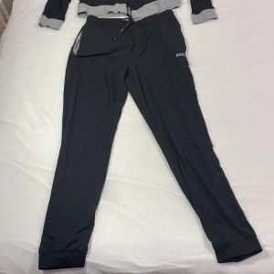 Dress från Boss - Säljer en exklusiv Hugo Boss mjukisdress i svart, som kombinerar lyxig komfort med stilren design. Setet består av en mjukisjacka och byxor, perfekt för både avslappnade tillfällen och när du vill ha en bekväm men ändå sofistikerad look. Hugo Boss är känt för att leverera premiumkläder med både funktion och stil, och denna dress är inget undantag.