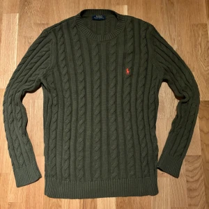 Ralph Lauren tröja - Säljer en snygg mörkgrön stickad tröja från Polo Ralph Lauren med kabelstickat mönster. Tröjan har en klassisk rund hals och långa ärmar. Den ikoniska loggan i rött finns broderad på bröstet. Perfekt för kyligare dagar! Tröjan passar M/L.