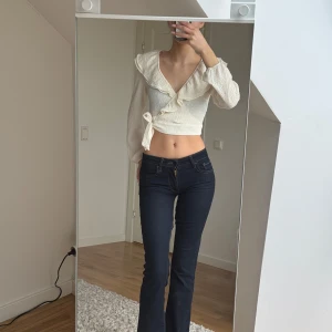 Vit omlottopp med volangdetaljer - Säljer en stilren vit omlottopp med volangdetaljer och knytning i sidan. Toppen har långa ärmar och en v-ringad design, perfekt för en chic look. Materialet är texturerat och ger en unik känsla. Passar perfekt till både jeans och kjol!