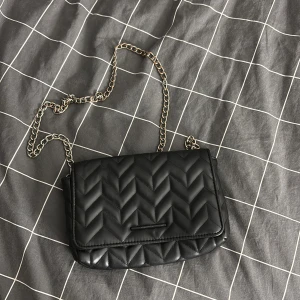 Svart axelväska med kedja - Snygg svart axelväska med en  kedjerem. Perfekt för att ge din outfit en stilfull touch. Väskan har en praktisk magnetknapp för enkel öppning och stängning. Helt okej kvalitet och köpt Fårn pull&bear tror inte den går att köpa längre