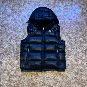 Svart dunväst från Moncler - Säljer en snygg svart dunväst från Moncler i mycket bra skick. Västen har en dragkedja framtill och praktiska fickor med dragkedjor. Perfekt för höst och vinter, och den har en cool, quiltad design. Passar både till vardags och lite finare tillfällen. Och den har ett hål i ryggen och en liten hål i fickan som inte syns utanför 