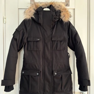 Svart parkajacka från Nobis - Säljer en svart parkajacka från Nobis modellen Cindy i mycket bra skick. Jackan har en avtagbar huva och flera praktiska fickor. Storlek: small Färg: svart Den är knappt använd och är i nyskick.   Säljer på grund av att jag har växt ur den.   Modellen har justerbar midja, vindtät, vattentät, ventilationsmöjligheter och är ”seam sealed”. 