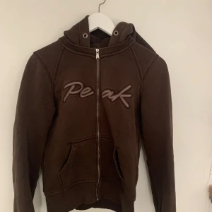 Brun hoodie från Peak - Säljer en brun hoodie från Peak Performance med dragkedja och huva. Den har ett broderat märke på framsidan! Jättesnygg!