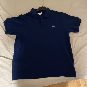 Mörkblå pikétröja från Lacoste - Säljer en snygg mörkblå pikétröja från Lacoste i mycket bra skick. Den har korta ärmar och den klassiska broderade krokodilen på bröstet. Perfekt för både vardag och lite finare tillfällen. Passar bra till jeans eller chinos!