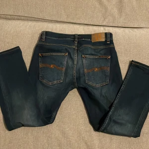 Mörkblå jeans från Nudie Jeans - Säljer ett par snygga mörkblå jeans från Nudie Jeans. De har en klassisk femficksdesign med orangea sömmar på bakfickorna. Jeansen är i bra skick och har en knappgylf. Perfekta för både vardag och fest! Storleken på jeansen är 31/32 men dom är uppsydda, skulle uppskatta längden runt 30!!
