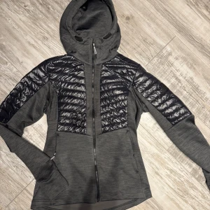 Grå jacka med svarta detaljer från Didriksons - Säljer en snygg grå jacka från Didriksons med svarta quiltade detaljer. Jackan har en dragkedja framtill och är perfekt för höst och vår. Den är i mycket bra skick och har en skön passform. Perfekt för dig som vill ha en stilren och funktionell jacka!