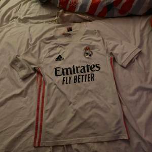 Säljer en vit Real Madrid fotbollströja från Adidas i storlek 164. Tröjan har korta ärmar och är i mycket bra skick. Den har klubbens emblem och sponsorlogga på framsidan. Perfekt för alla fotbollsfans! ⚽Tröjan passar XS och S väldigt bra också👍