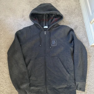Grå hoodie från Burberry - Säljer en snygg grå hoodie från Burberry i bra skick. Den har en dragkedja framtill och en klassisk logga på bröstet. Insidan av huvan har ett stilrent rutigt mönster. Perfekt för höst och vinter! Har använt den en gång,det är för stor för mig .köpt den förra året 