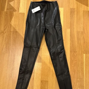 Svarta skinnleggings - Snygga svarta skinnleggings i nytt skick. De är högmidjade och har en tight passform som ger en cool look. Perfekta för både fest och vardag. Materialet är syntetiskt skinn som ger en glansig finish.