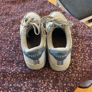 Nike Air Force i vitt och blått - Säljer ett par klassiska Nike Air Force sneakers i vitt med blå detaljer. Skorna har snörning och är i acceptabelt skick med lite slitage. Perfekta för vardagsbruk och ger en sportig look. Storlek 38,5 bra passform. Köptes för 1200 kr