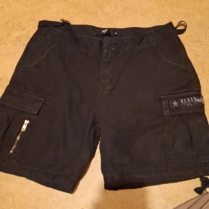 Svarta cargo shorts med dragkedja - Säljer ett par svarta cargo shorts från Black Star. De har praktiska fickor med dragkedja och knappar, perfekt för sommarens äventyr. Shorts i loose fit med justerbara remmar i midjan för bästa passform. Perfekta för både vardag och utflykter!