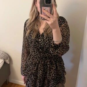 Leopardmönstrad klänning - Säljer en superfin leopardmönstrad klänning med v-ringning och knappar framtill. Den har långa ärmar och en volangdetalj i nederkant. Perfekt för både vardag och fest! 🐆✨