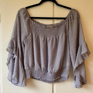 Ljuslila/grå blus med volangärmar - Säljer en superfin ljuslila blus med volangärmar och smockdetaljer vid hals och midja. Blusen har ett diskret prickigt mönster och är perfekt för både vardag och fest. Off-shouldermodell.