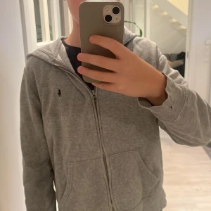 Grå hoodie från Ralph Lauren - Säljer en grå hoodie från Ralph Lauren med dragkedja och det klassiska logotypen på bröstet. Den har långa ärmar och en liten skada på ena ärmen. Perfekt för en avslappnad stil. Hör av dig vid intresse!