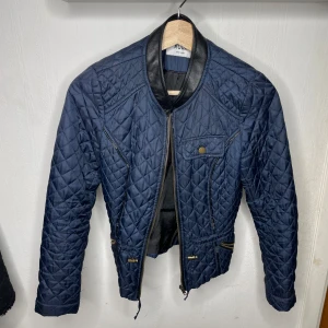 Mörkblå quiltad jacka från Mudd - Säljer en snygg mörkblå quiltad jacka från Mudd. Jackan har en dragkedja framtill och en cool krage i skinn. Perfekt för höst och vår med sin stilrena design och bekväma passform. Den har långa ärmar och en ficka med knappdetalj på bröstet.