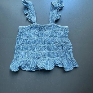 Blå randig topp med volangdetaljer - Söt blå randig topp med volangdetaljer och smockad design. Perfekt för sommardagar! Den har breda axelband och en luftig passform som gör den bekväm att bära. Passar bra till både jeans och kjol för en avslappnad look.