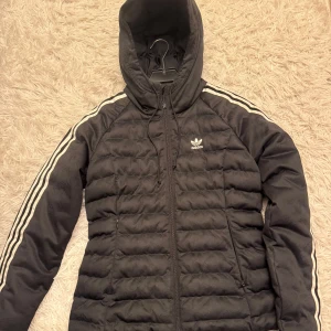 Svart puffer slim jacka från Adidas - Säljer en snygg svart pufferjacka från Adidas med klassiska vita ränder längs ärmarna. Jackan har en huva och dragkedja framtill, perfekt för kyliga dagar. Den är i mycket bra skick och passar både till vardags och sportiga aktiviteter. Har inte tagit på mig den mer än 5 gånger bara inte min stil så den är ny.
