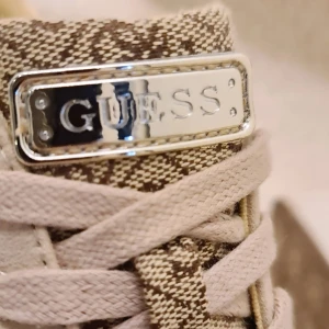 Beige sneakers från Guess - Snygga beige sneakers med kilklack från Guess med ett coolt mönster och metallic-detaljer. Skorna har snörning och en bekväm sula, perfekt för vardagsbruk. De är i bra skick och har en stilren design som passar till det mesta.