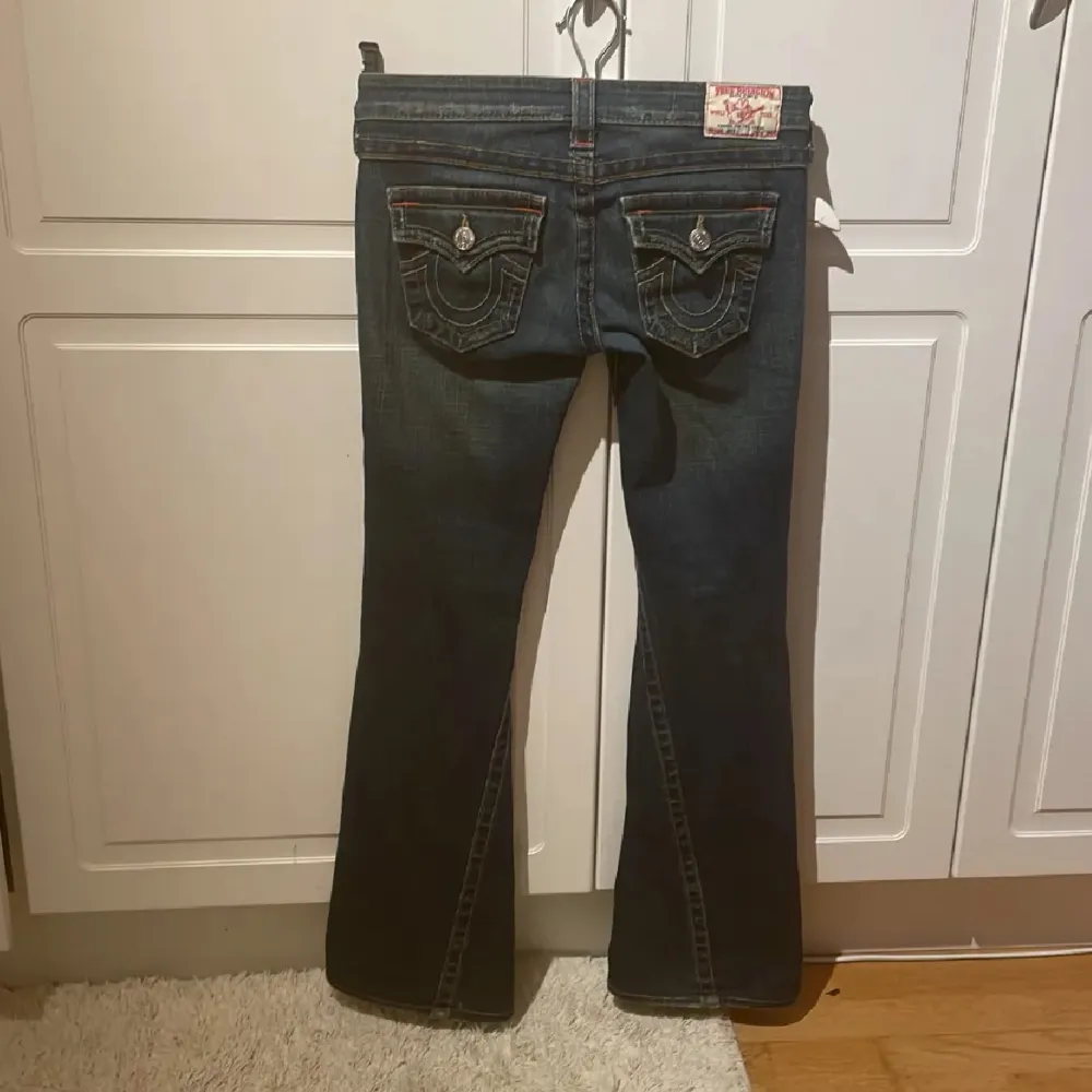 Säljer ett par snygga mörkblå bootcut jeans från True Religion. De har en låg midja och coola slitningar på framsidan. Bakfickorna har snygga broderade detaljer och knappar. Perfekta för en avslappnad men stilren look!. Jeans & Byxor.