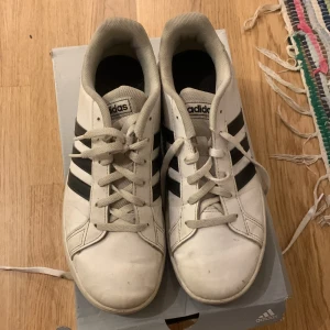 Adidas sneakers i vitt och svart - Säljer ett par klassiska Adidas sneakers i vitt med svarta ränder. Skorna har snörning och är i bra skick. Perfekta för vardagsbruk och ger en sportig look. Tillverkade i Indonesien.