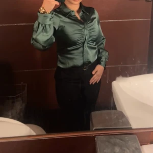 Grön satinblus - Snygg grön satinblus med långa ärmar och knappar framtill. Perfekt för en festlig kväll eller en elegant middag. Blusen har en v-ringad design och en åtsittande passform som framhäver figuren. Den glänsande satinfinishen ger en lyxig känsla.