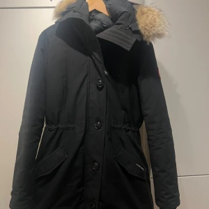 Svart parkajacka med pälsdetalj - Säljer en svart parkajacka från Canada Goose i mycket bra skick. Jackan har äkta päls på huvan och knappar framtill. Perfekt för kalla vinterdagar! Den har en snygg och stilren design med en broderad logga på ärmen. Den är knappt använd och som en ny. Köpt för 13000 kr just nu ligger den runt 15-17 tusen 