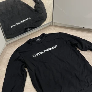 Emporio Armani tröa (sweatshirt) - Säljer en stilren svart tröja från Emporio Armani i storlek L. Tröjan har en klassisk design med långärmade ärmar och det ikoniska Emporio Armani-logotypen tryckt i vitt på bröstet. Perfekt för både vardag och festliga tillfällen. Passar bra till jeans eller chinos för en avslappnad men ändå elegant look.