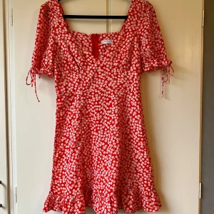 Röd blommig klänning - Superfin röd klänning med vita blommor. Den har korta ärmar med knytband och en söt volangkant längst ner. Klänningen är i ett lätt och luftigt material, vilket gör den idealisk för varma dagar. Passar både till vardags och festliga tillfällen.